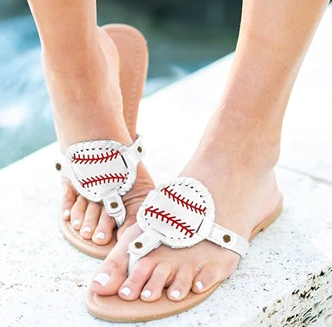 White sandals with baseball design on toes worn by a person sitting on a rock by water.