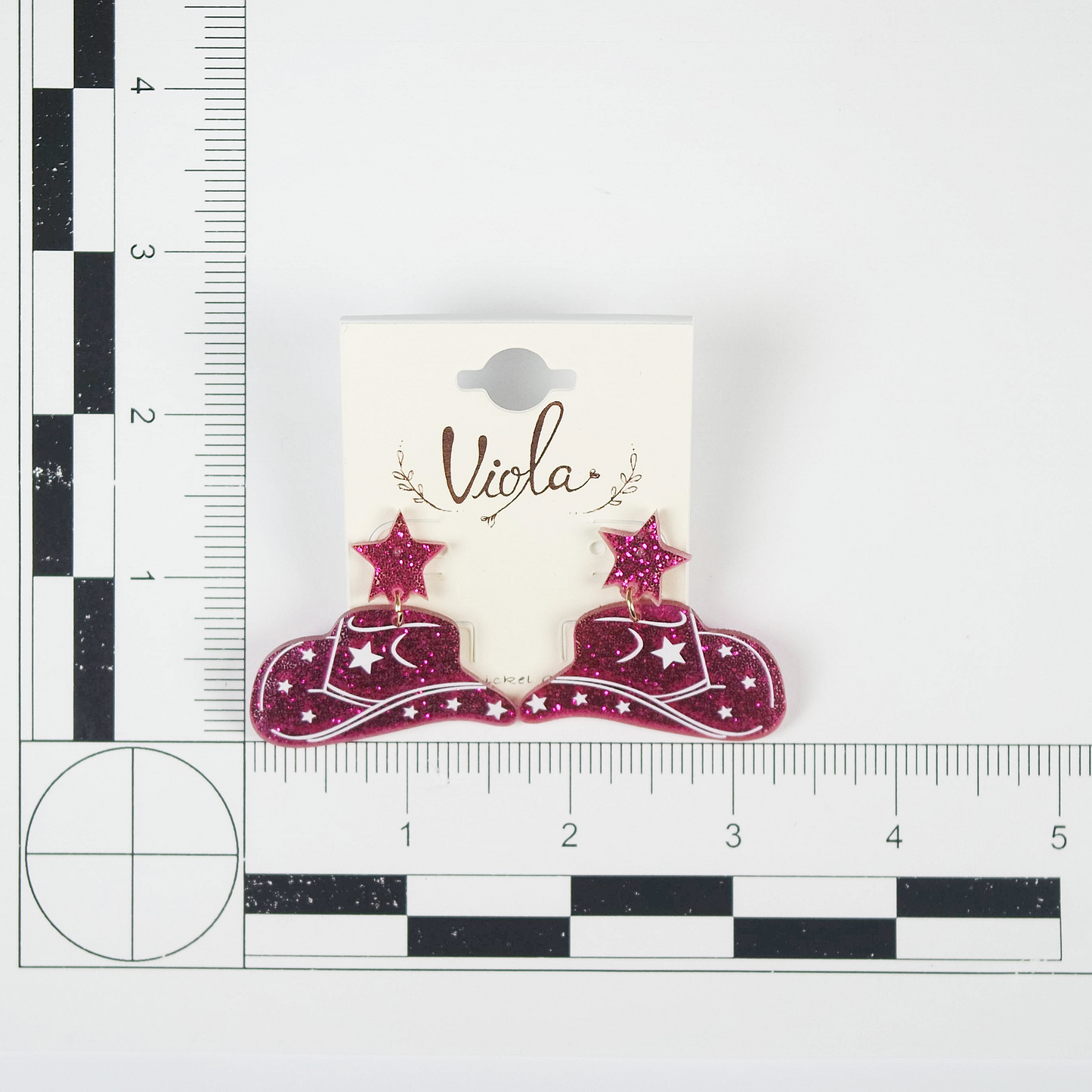 Card with pink cowboy hat and star earrings on a white background, next to a ruler for scale.