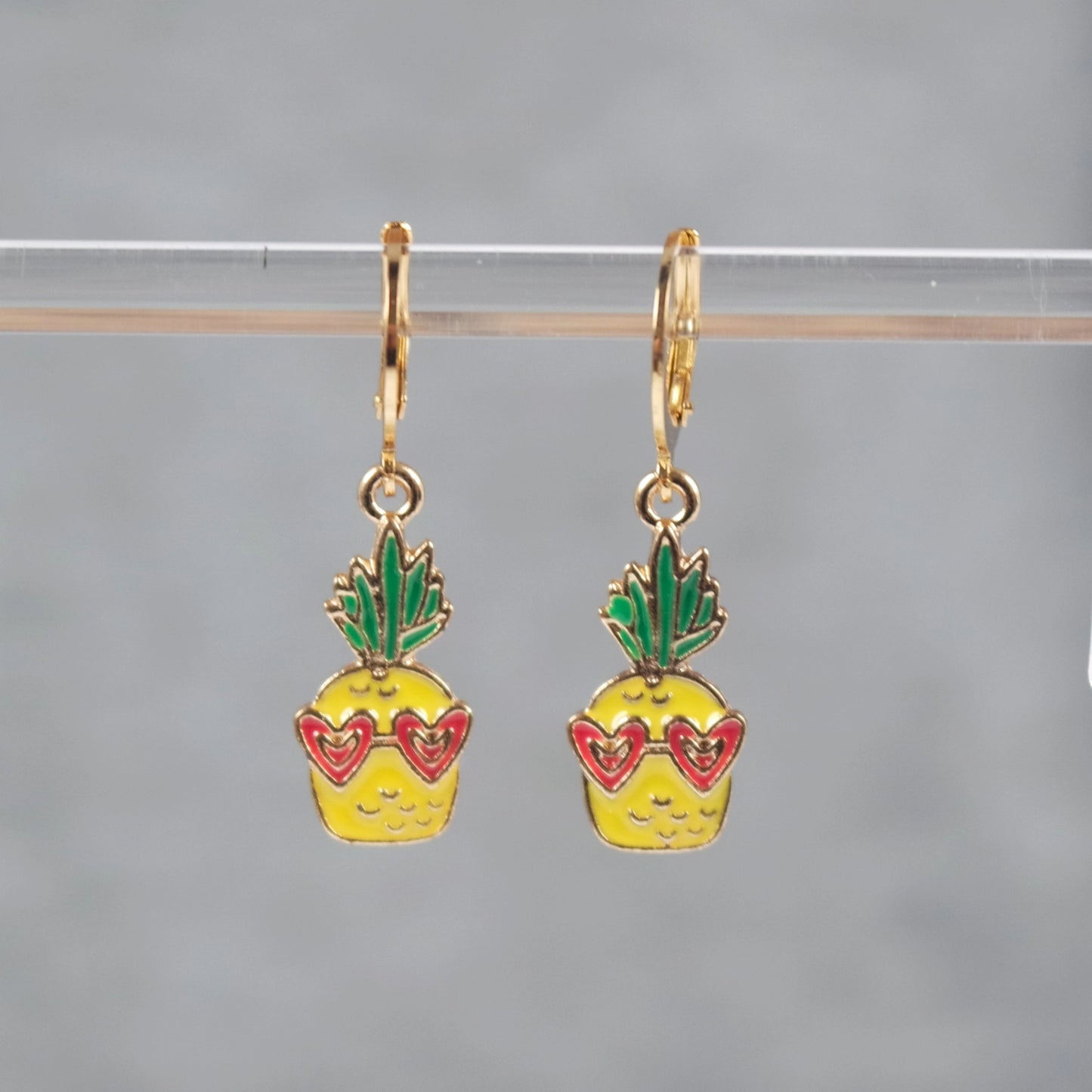 A pair of dangle earrings featuring a pineapple with a pink heart-shaped sunglasses design, attached to gold-colored hoop earrings.