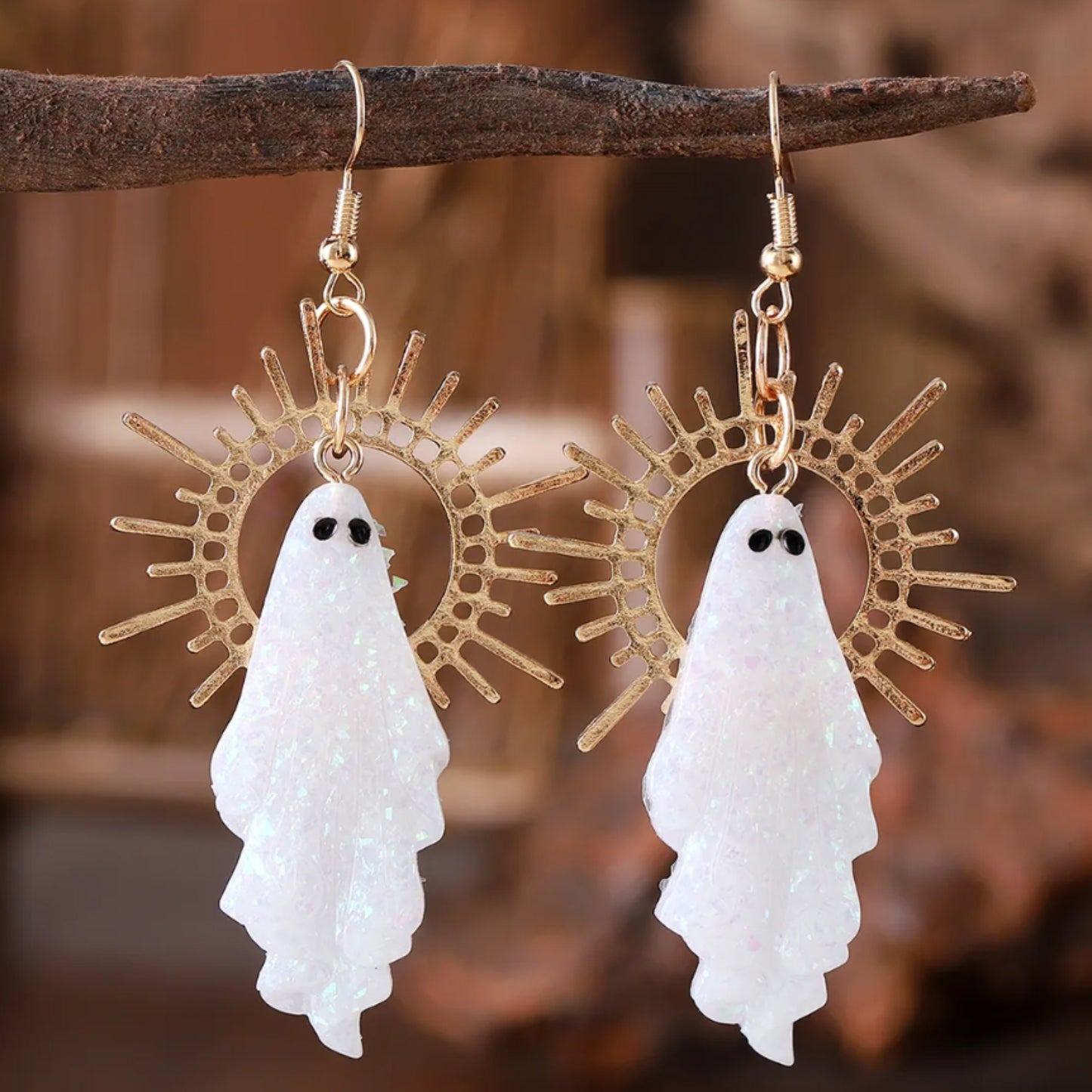 Ghost-shaped earrings with gold sunburst designs on a wooden branch.