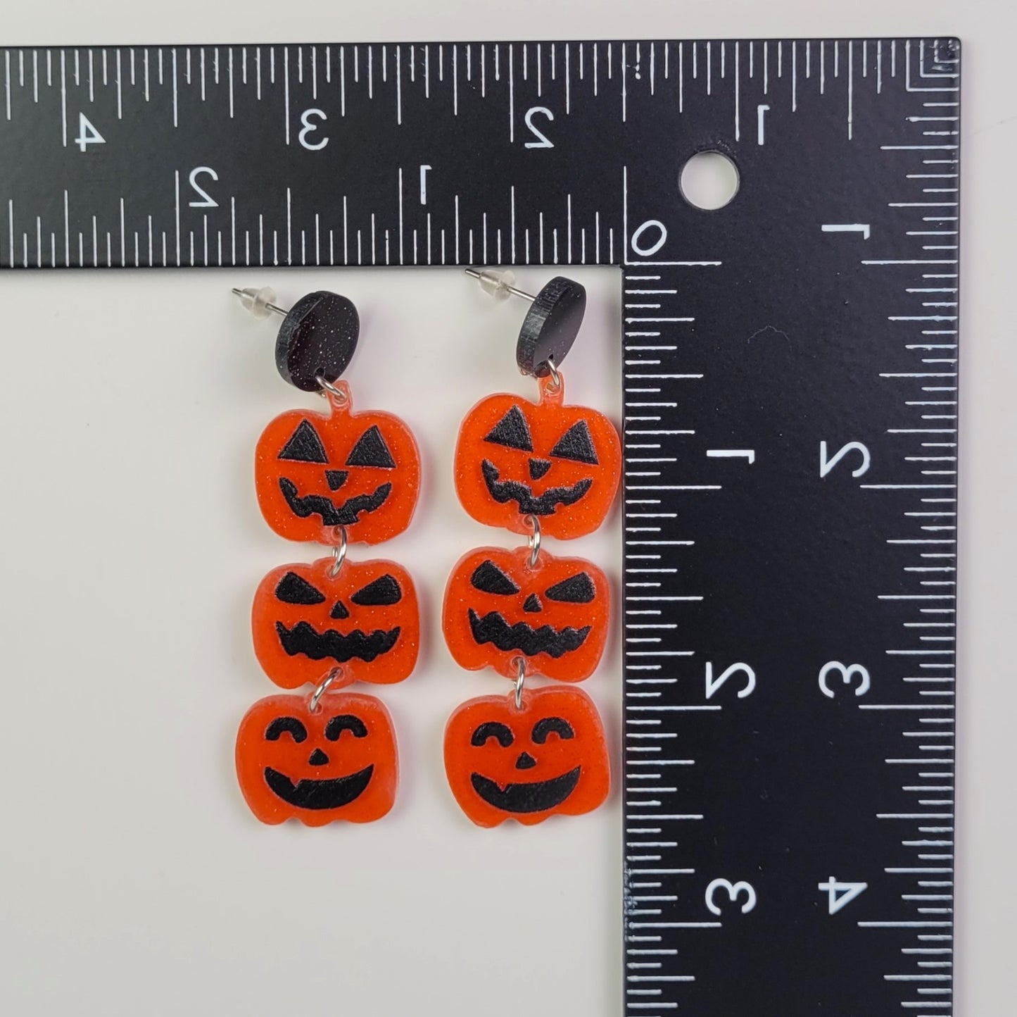 Halloween-themed earrings resembling jack-o'-lanterns on a white background with a ruler for scale.