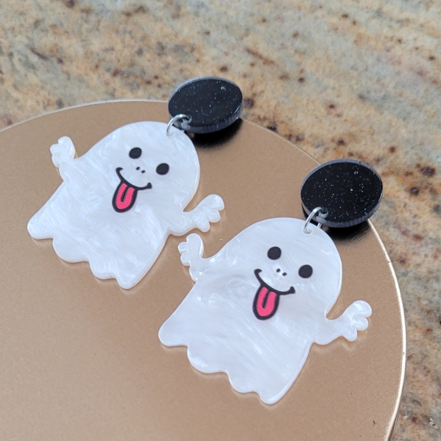 Pair of ghost-shaped earrings on a round metallic surface.