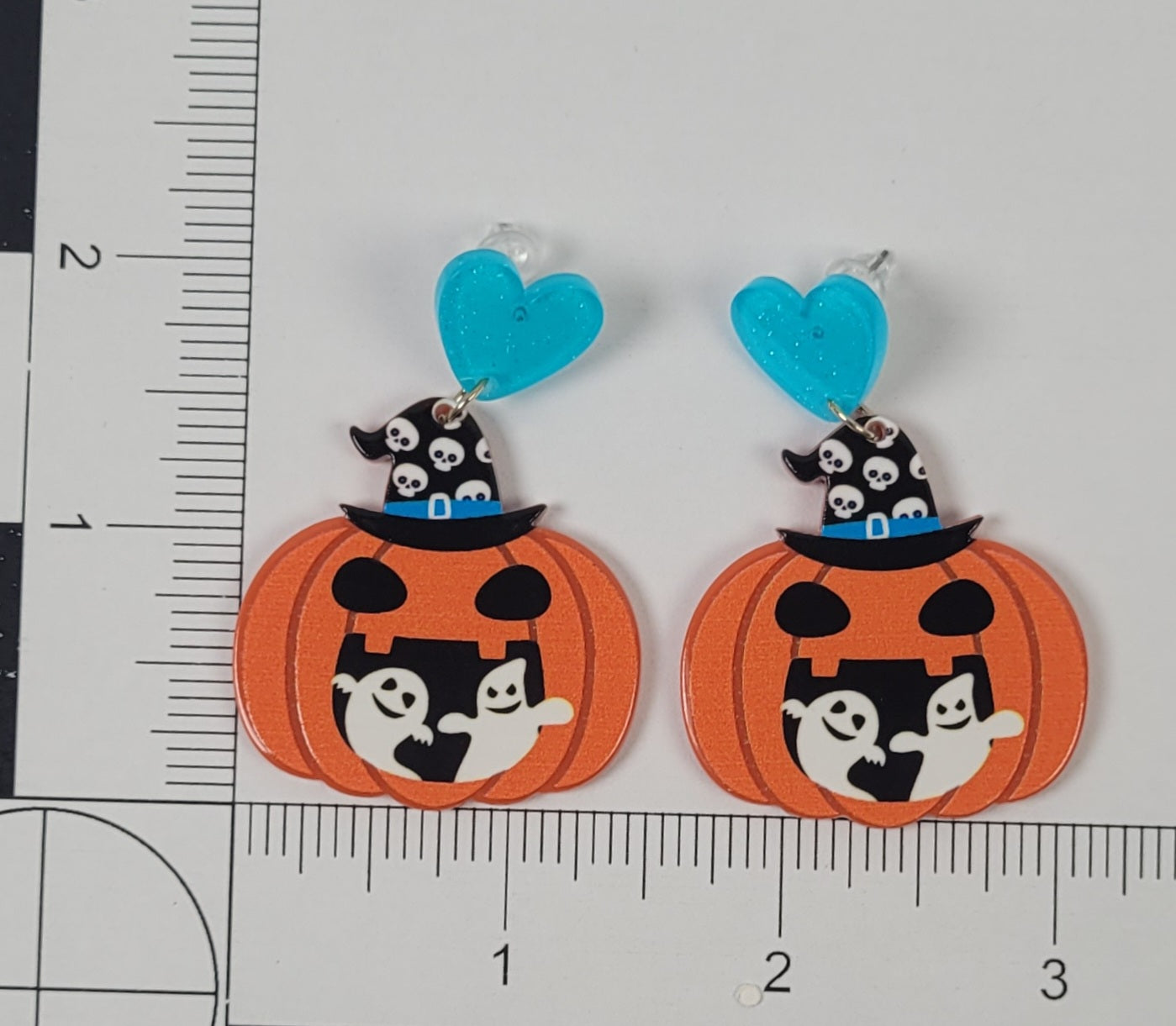 Halloween-themed pumpkin earrings with ghost and witch designs on a white background with a ruler for scale.