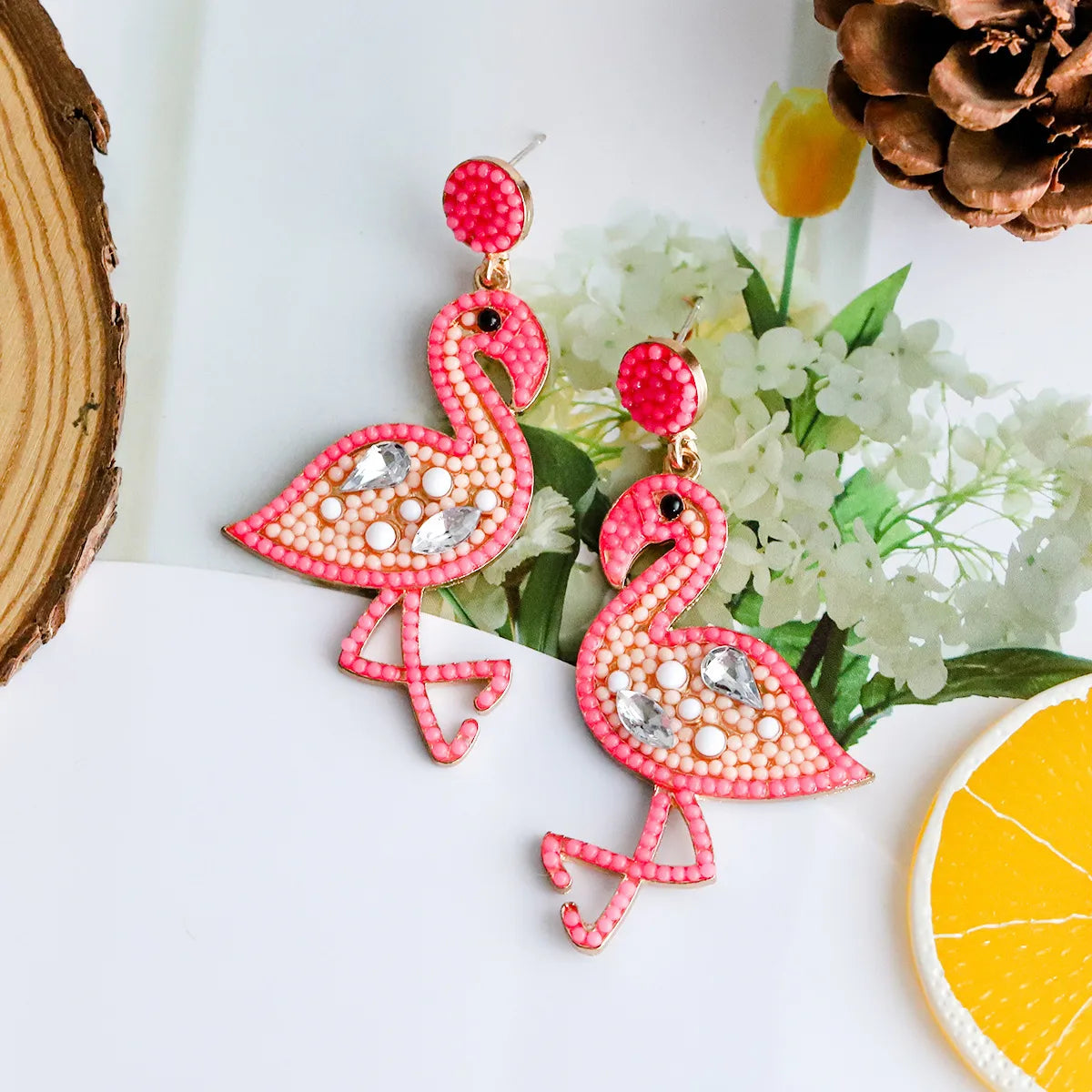 A pair of pink flamingo-shaped earrings adorned with multi-colored seeds beads, displayed against a beach-themed background with a slice of orange and a pine cone.