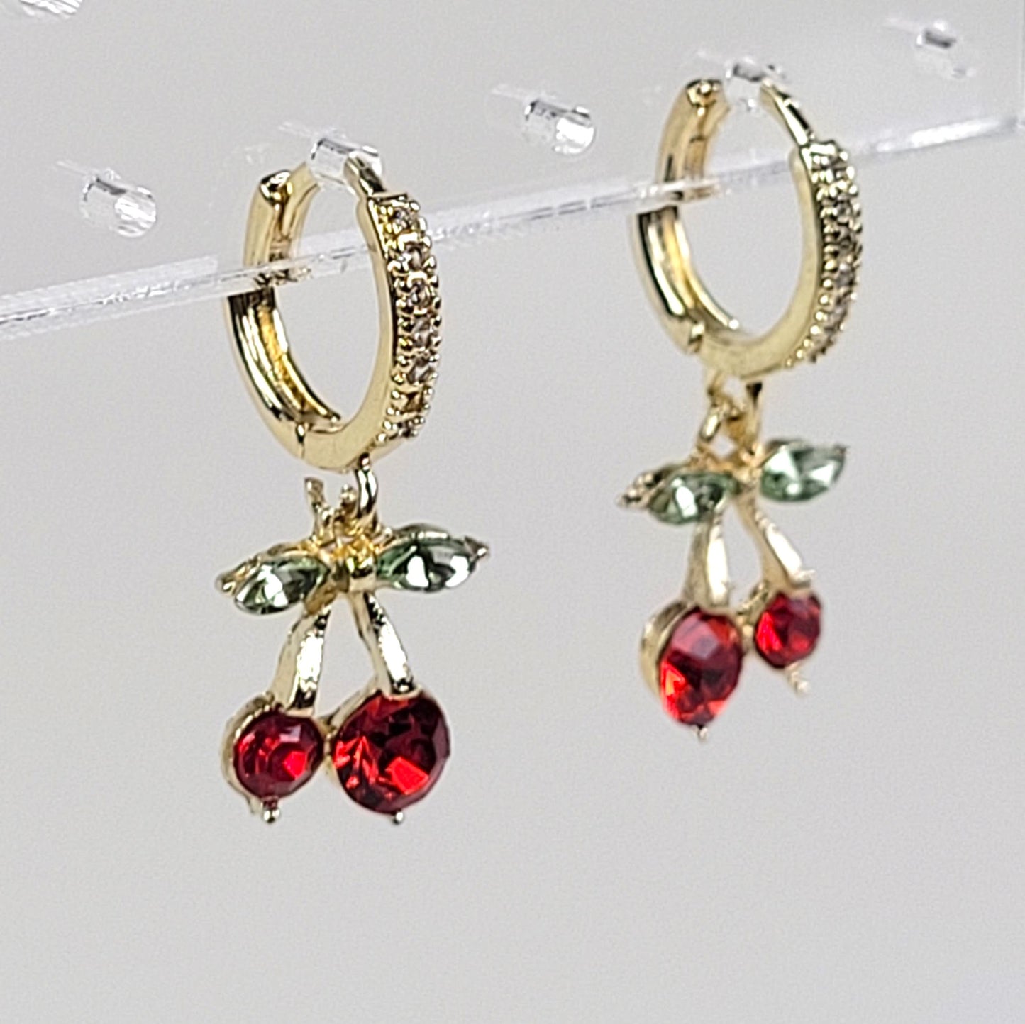 Gold hoop earrings with red and green gemstone charms on a clear stand.