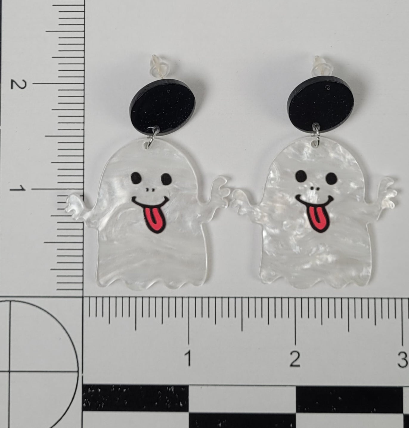 Ghost-shaped earrings with black eyes and red tongues on a ruler for scale.