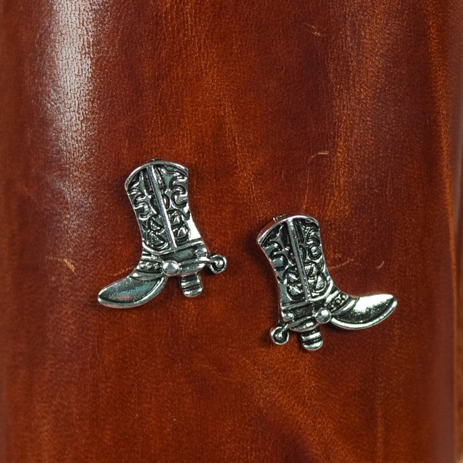 Silver cowboy boot earrings on a brown leather surface