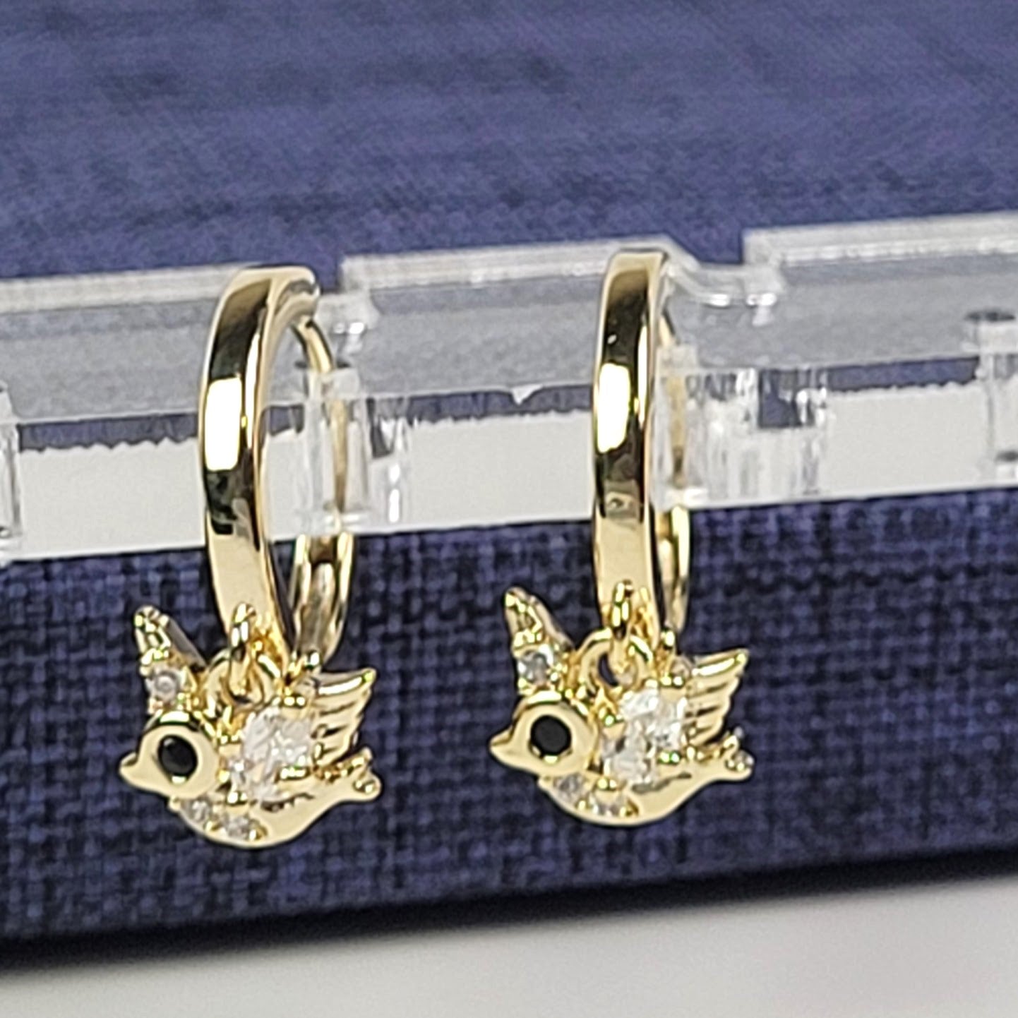Gold bird-shaped earrings on a clear stand with a blue fabric background