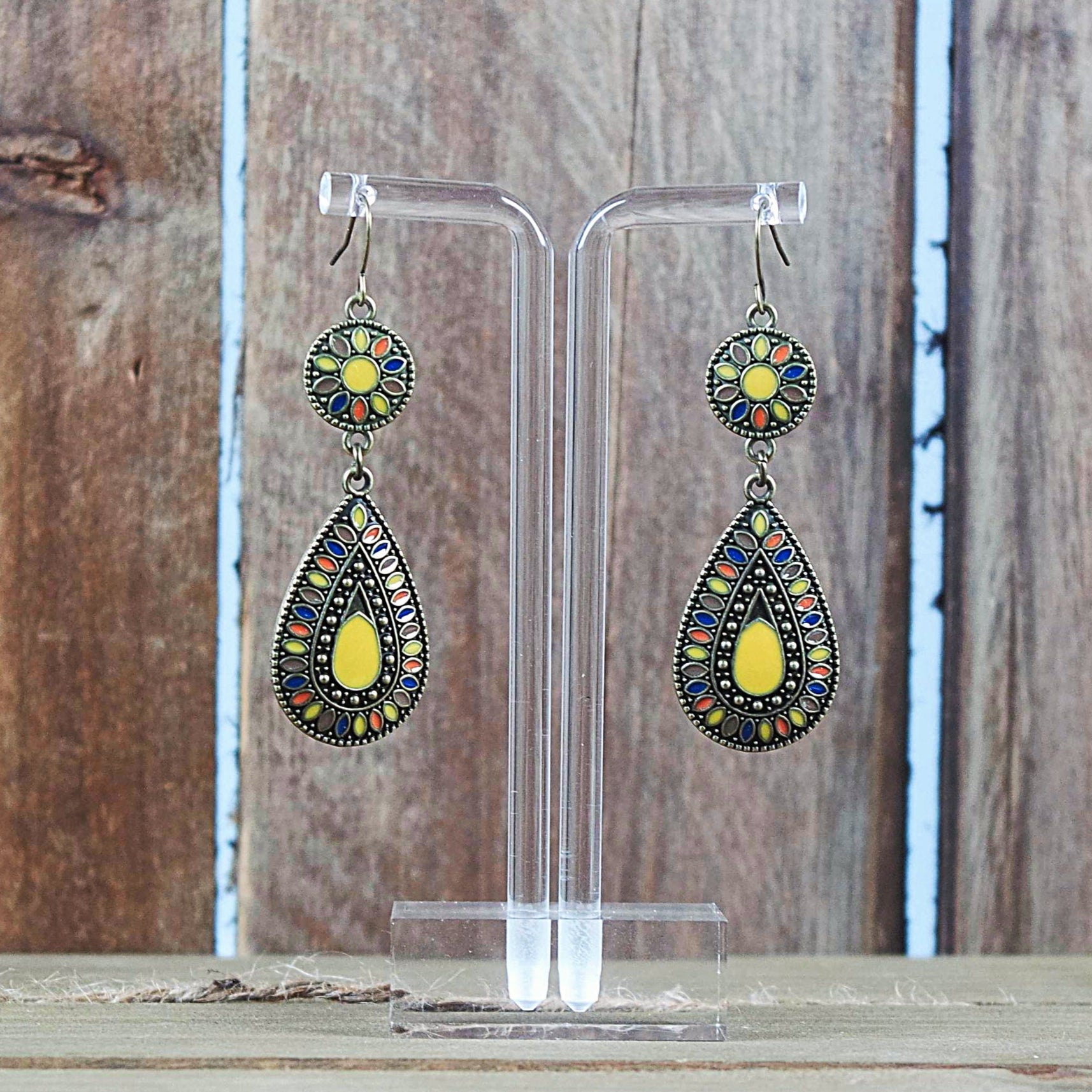 A pair of bohemian style drop earrings with antique brass finish and multicolor yellow enamel detailing, displayed against a wooden background.