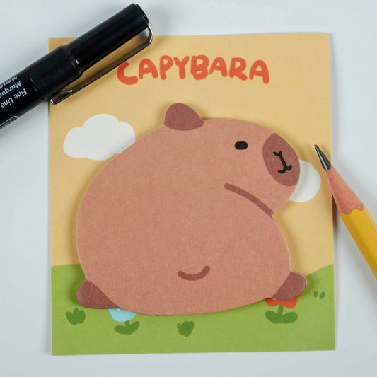 A cartoon image of a cute capybara on a sticky note pad with a pen and a pencil lying next to it.