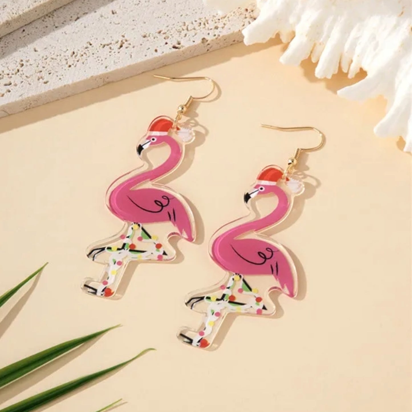 Pink flamingo earrings on a beige surface with coral and green leaves.