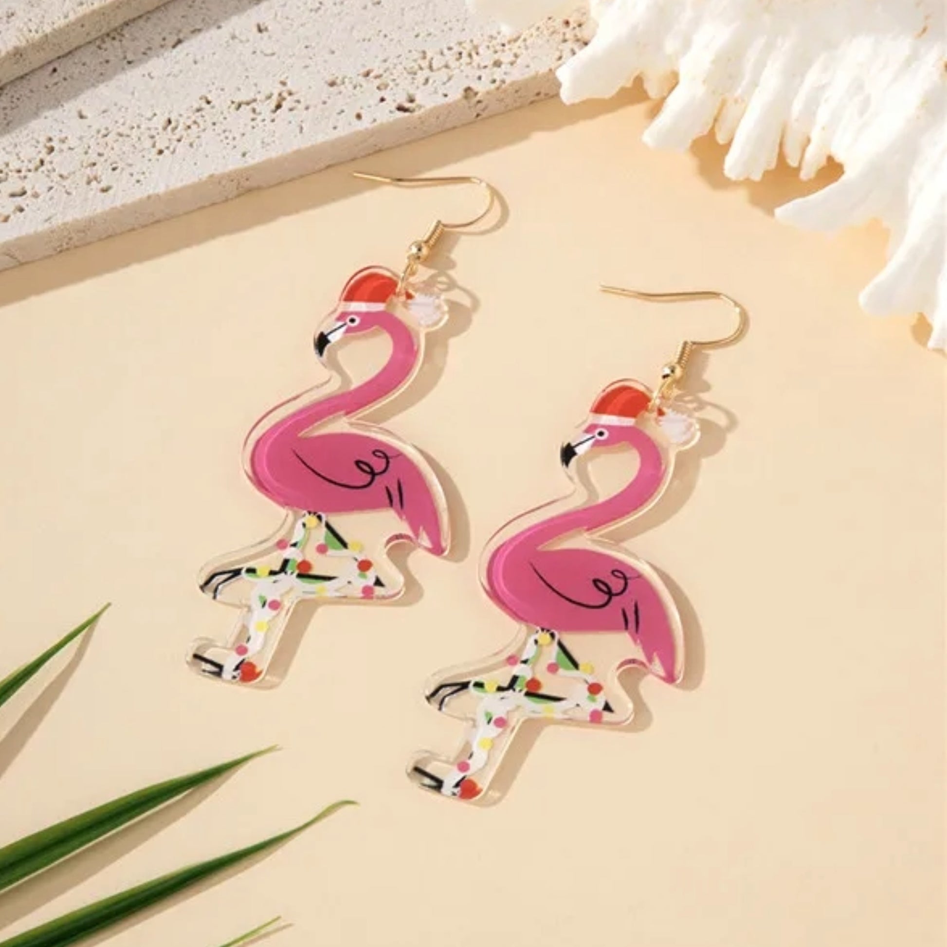 Pink flamingo earrings on a beige surface with coral and green leaves.
