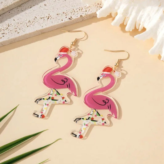 Pink flamingo earrings on a beige surface with coral and green leaves.