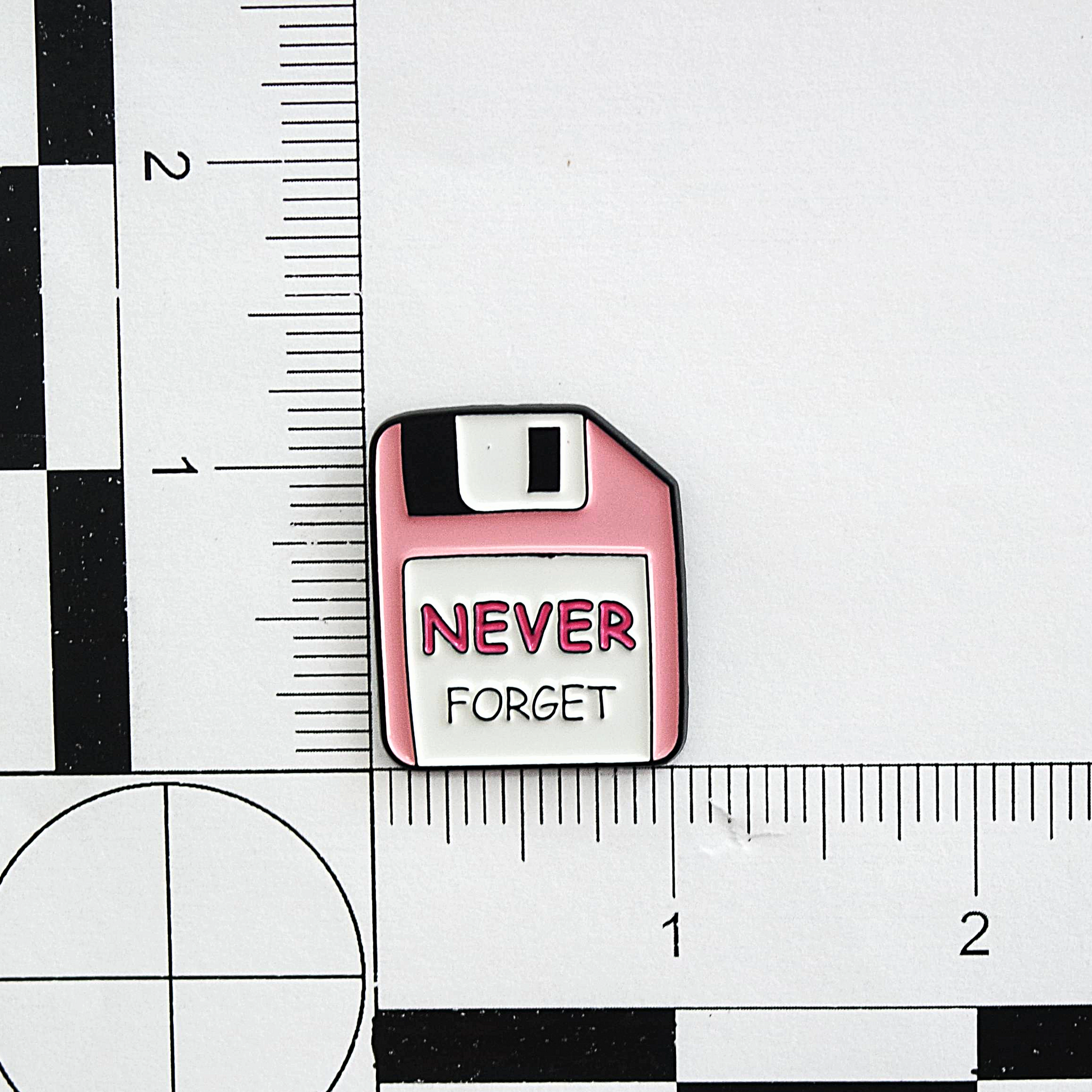 Pink floppy disk-shaped enamel pin with 'NEVER FORGET' text on a white background with ruler for scale.