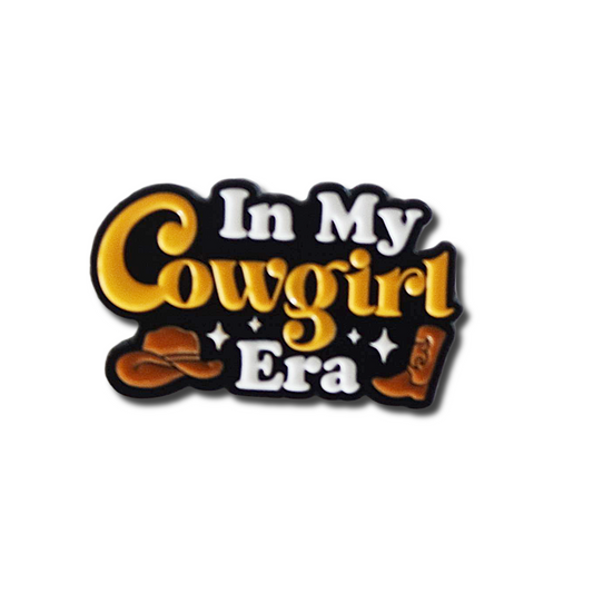 Decorative enamel pin with 'In My Cowgirl Era' text and cowboy hat and boot graphics on a white background.
