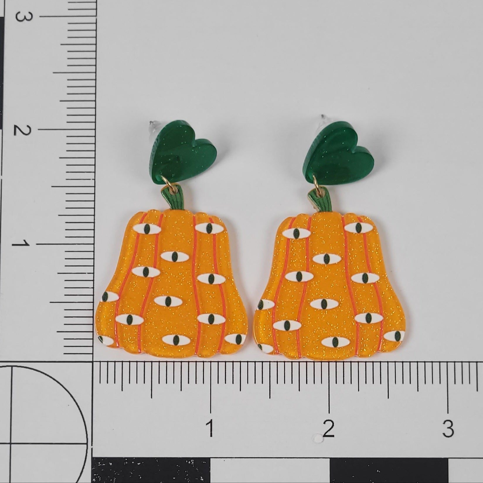 Pumpkin-shaped earrings with green leaves on a white background with a ruler for scale.