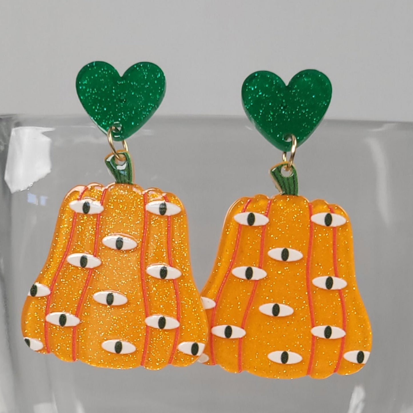 Pair of earrings with orange pumpkin-like design  with evil eyes and green heart-shaped tops on a clear background