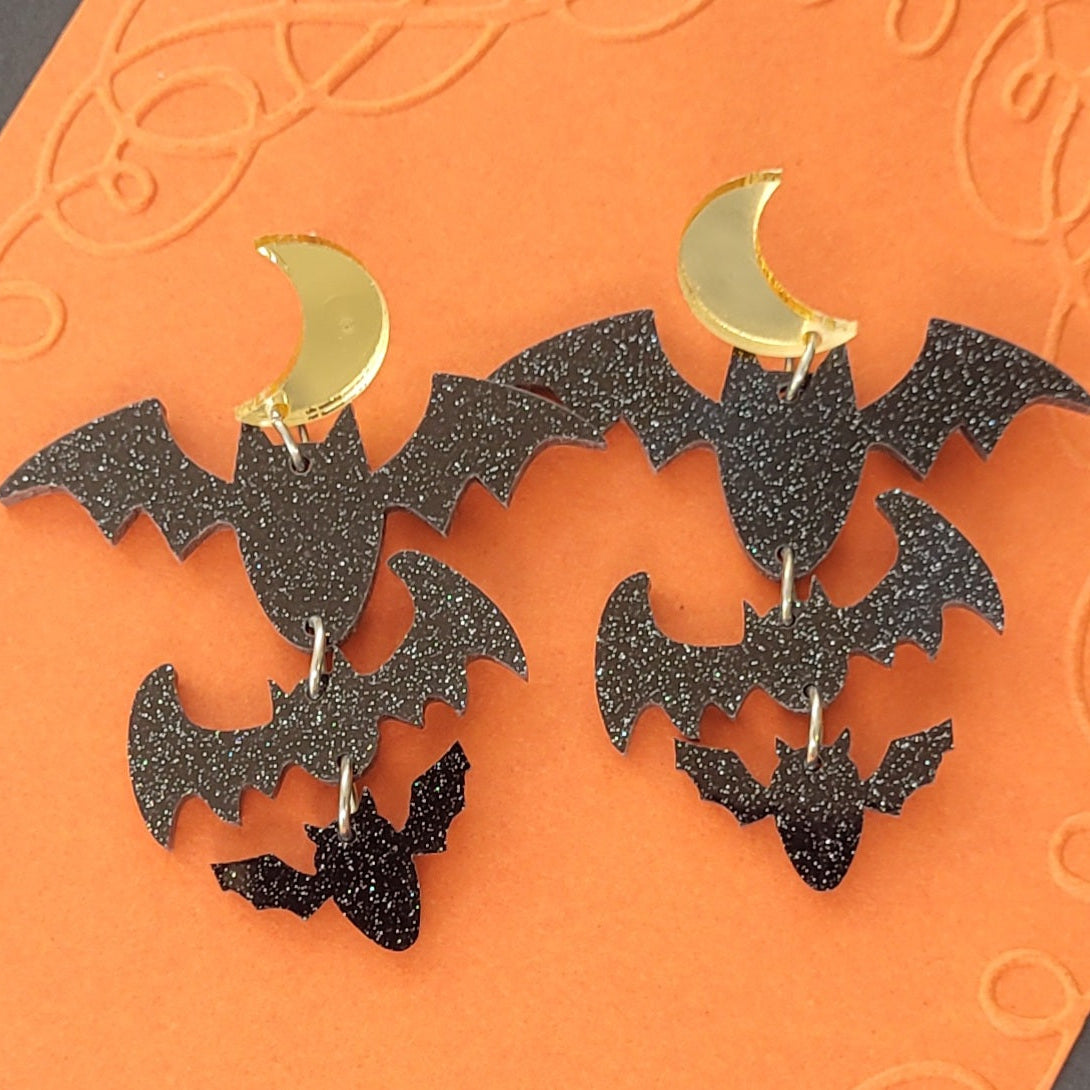 Black glitter bat earrings with gold crescent moons on an orange background