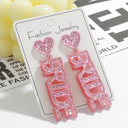 A pair of pink glittering acrylic earrings with the word 'Bride' written in letters and a heart shape at the stud, displayed on a white card with 'Fashion Jewelry' written at the top.