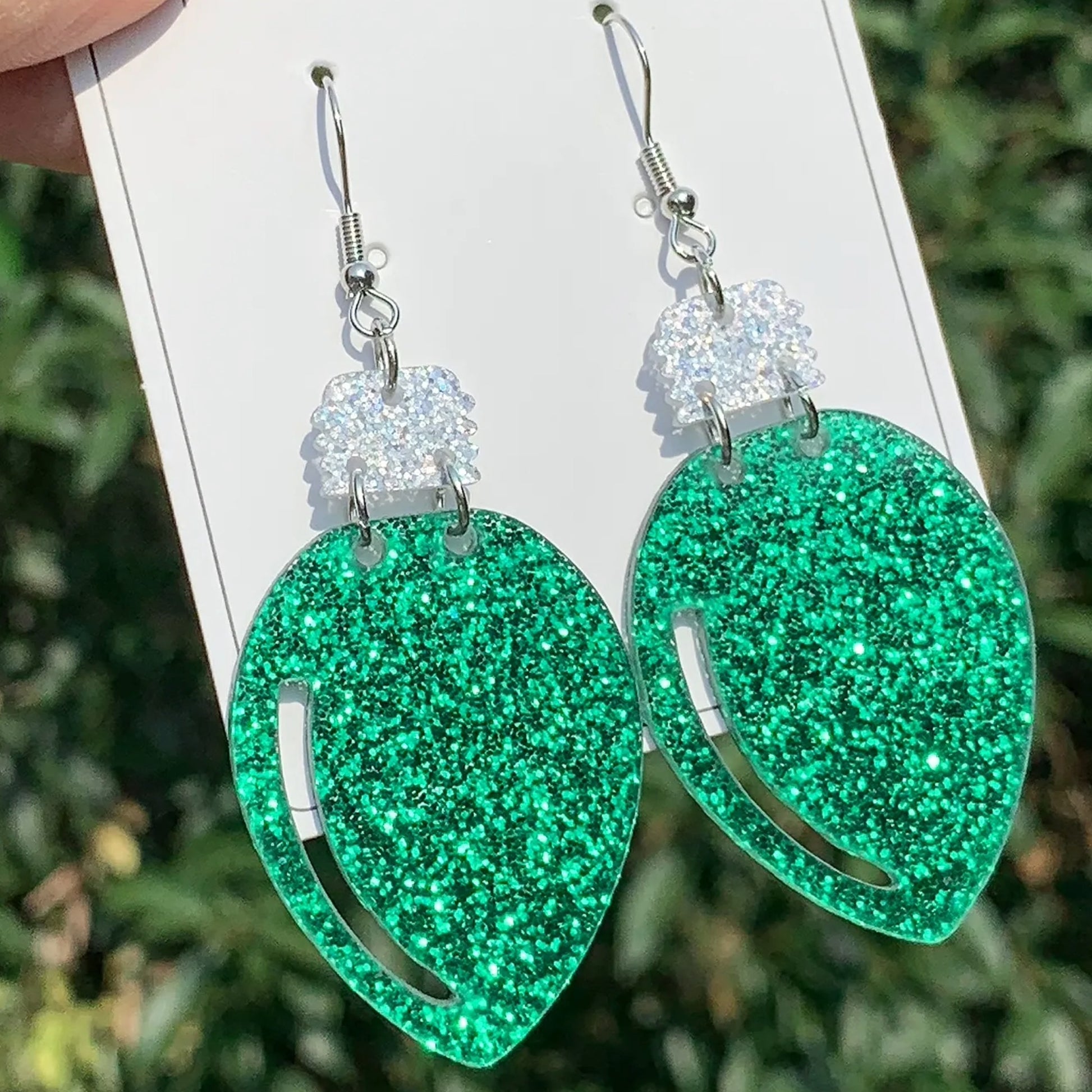 Green glittery earrings on a white card with a blurred green background