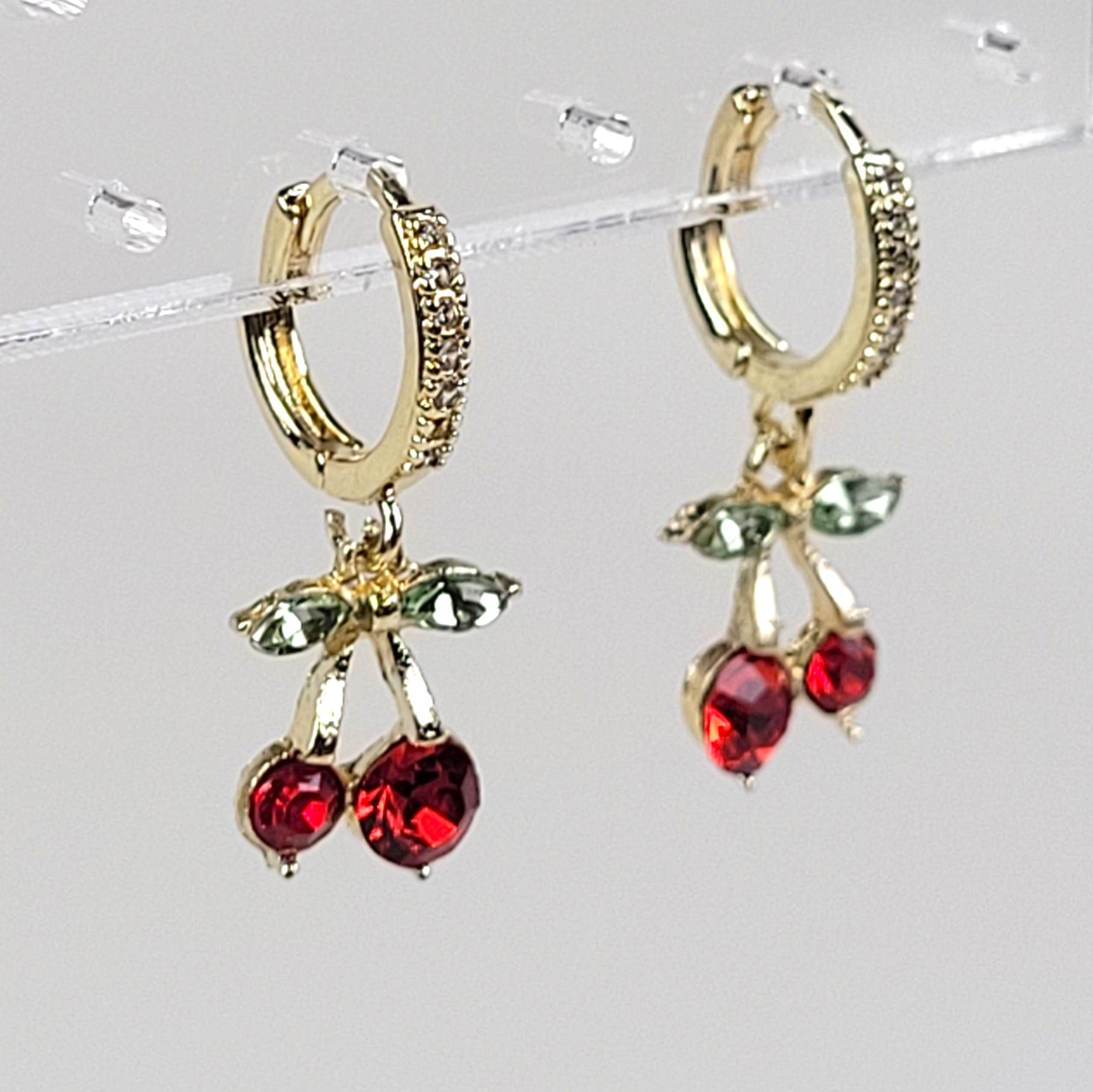 Gold hoop earrings with red and green gemstone charms on a clear stand.