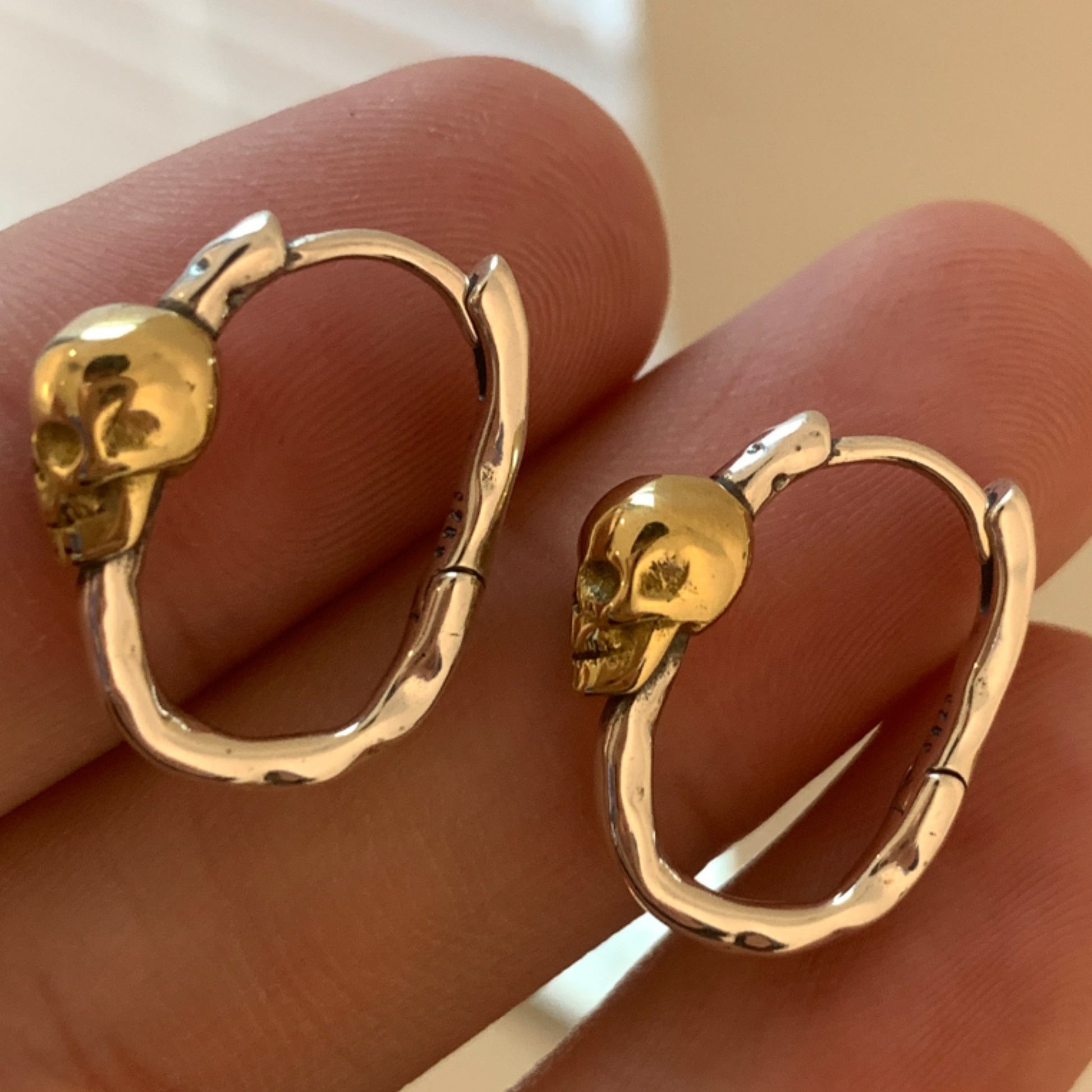 Gold skull hoop earrings on a hand against a neutral background