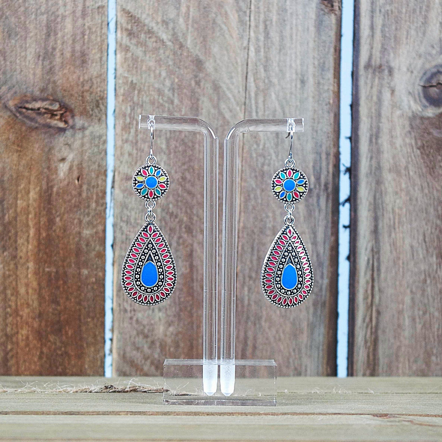 A pair of silver-tone finish dangle earrings with multicolor blue and red enamel, displayed against a wooden background.