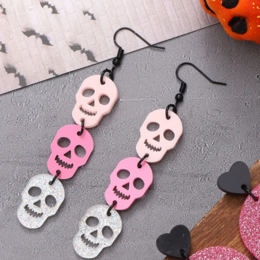 Skull-shaped earrings in pink, silver, and black on a gray surface with Halloween-themed decorations.