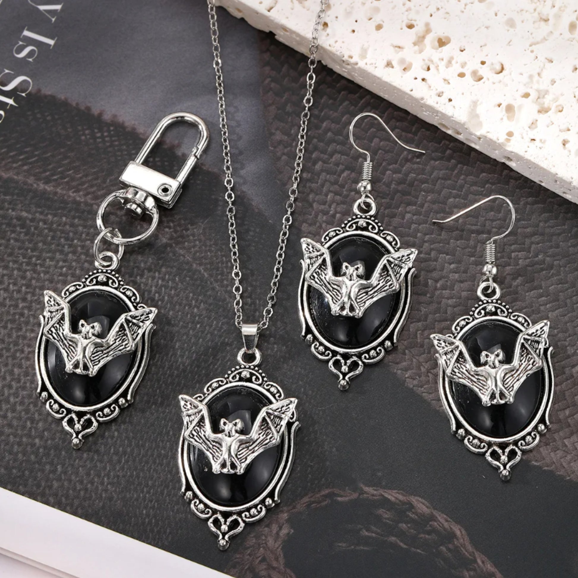 Jewelry set with bat designs on black stones, including a necklace, pendant, and earrings, on a textured surface.